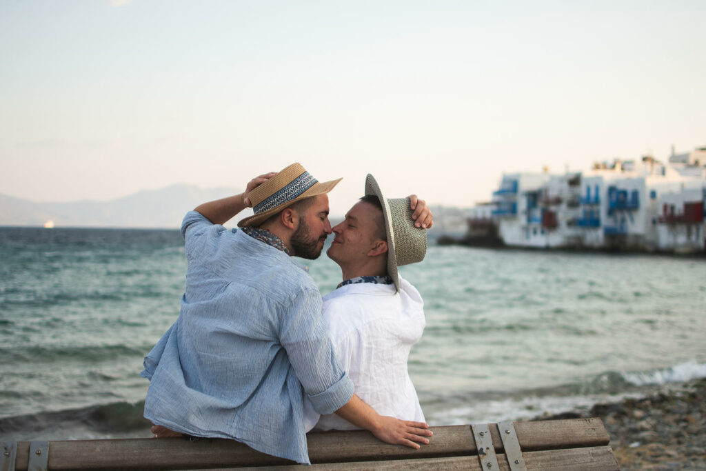 Male couple in straw hats sitting on a bench in front of Little Venice in Mykonos