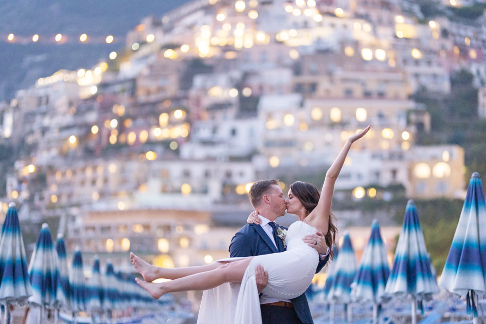 The groom hold the bride without her shoes for photo in front of the hill by sunset