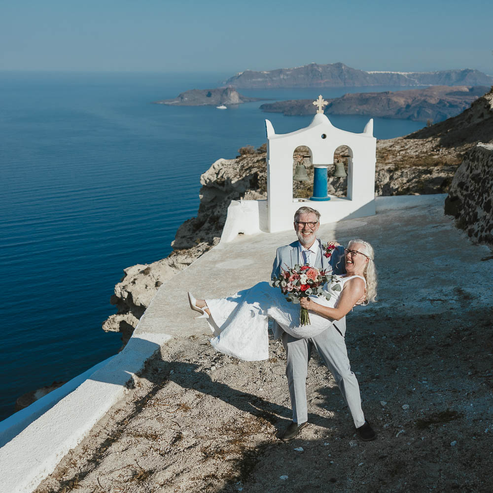 Vows renewal in Santorini – 017 Just like 25 years ago, the husband carries his wife in his arms with the Santorini landscape as background