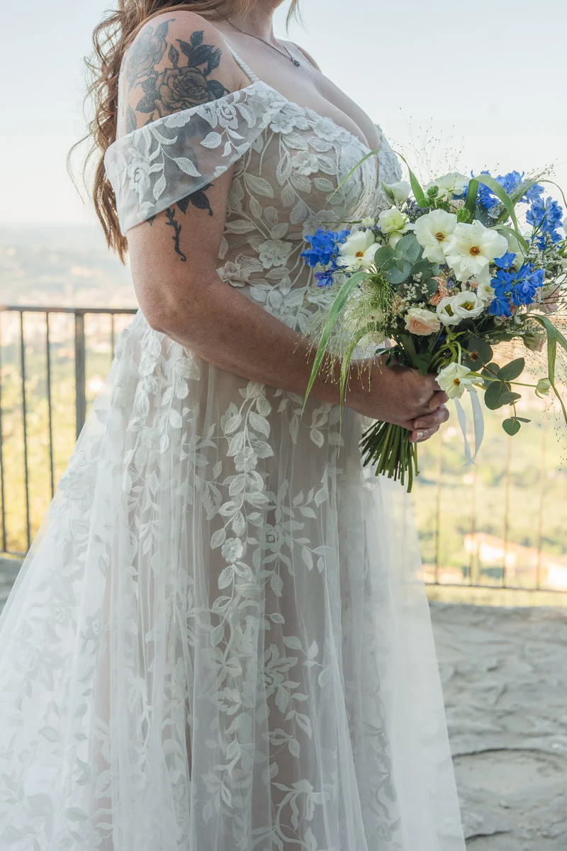 elopment at Fiesole, Tuscany-05 Close-up of bride's lace off-shoulder wedding dress and wildflower bridal bouquet with blue delphiniums and white blooms, Fiesole terrace