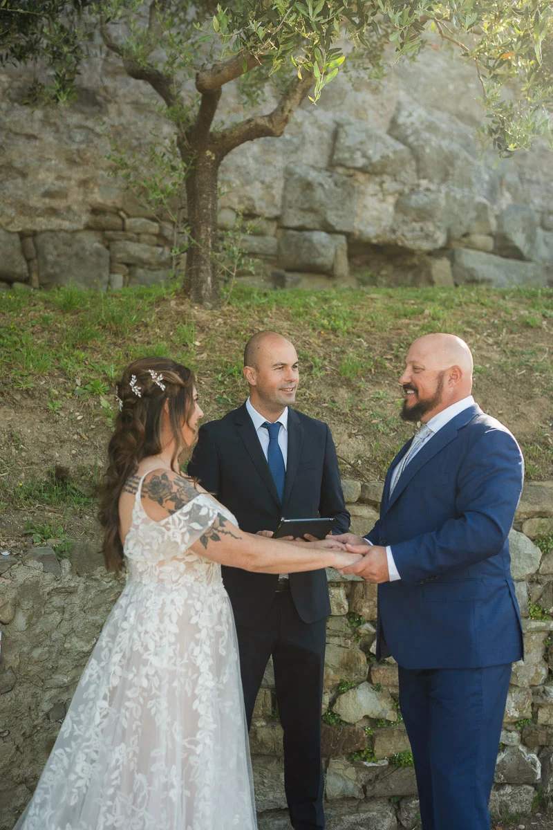 elopment at Fiesole, Tuscany-16 Bride and groom holding hands during their intimate elopement ceremony in Fiesole, with an olive tree and ancient stone wall behind them