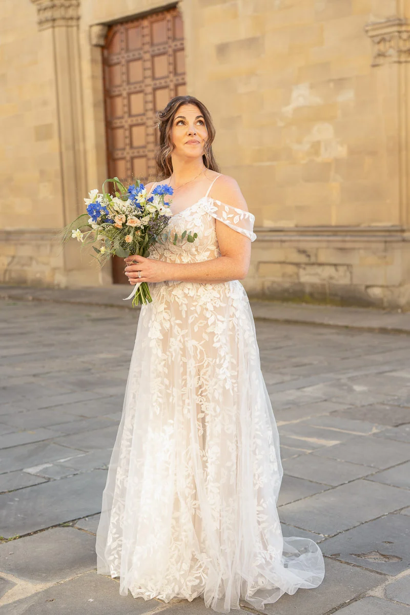 elopment at Fiesole, Tuscany-23 Bride in off-shoulder lace wedding gown holding a wildflower bouquet on a sunlit stone square in Fiesole, Tuscany