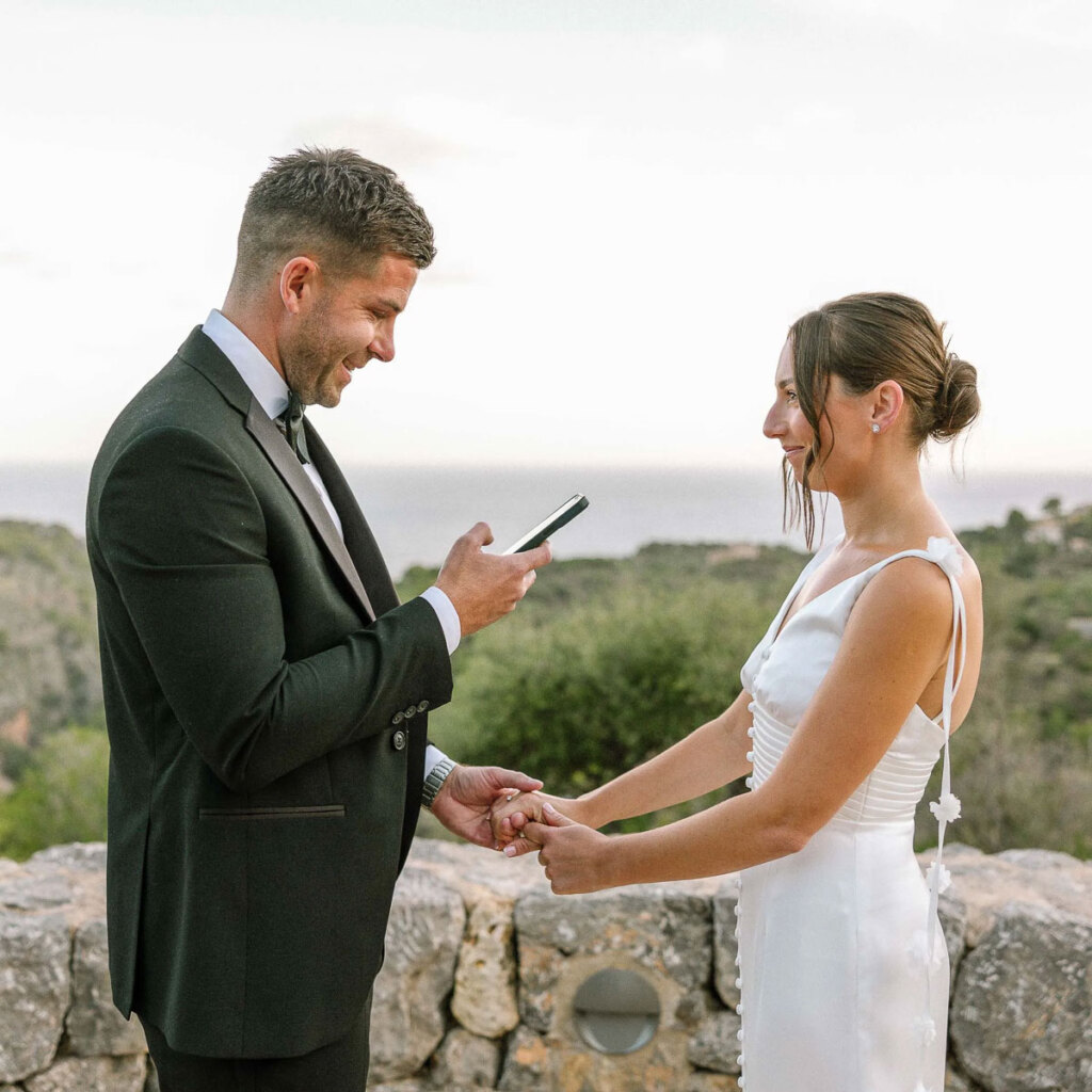 A groom reading his wedding vows from a phone while holding the bride's hand during an intimate sunset elopement ceremony on a rocky cliffside in Mallorca, Spain.